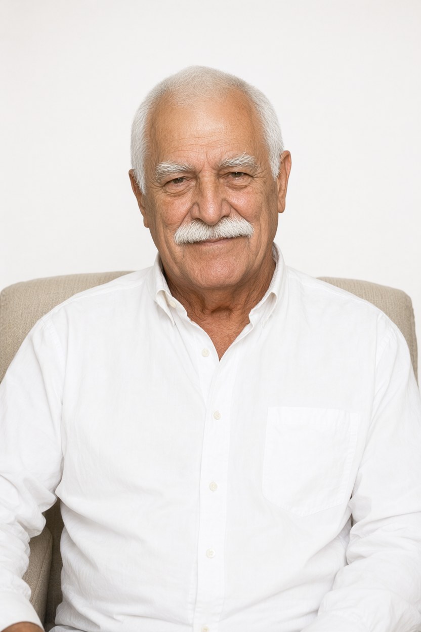 Waist-up medium shot in a plain white shirt with no cardigan, seated in the armchair, hair freshly combed, soft even daylight, looking just past the camera.